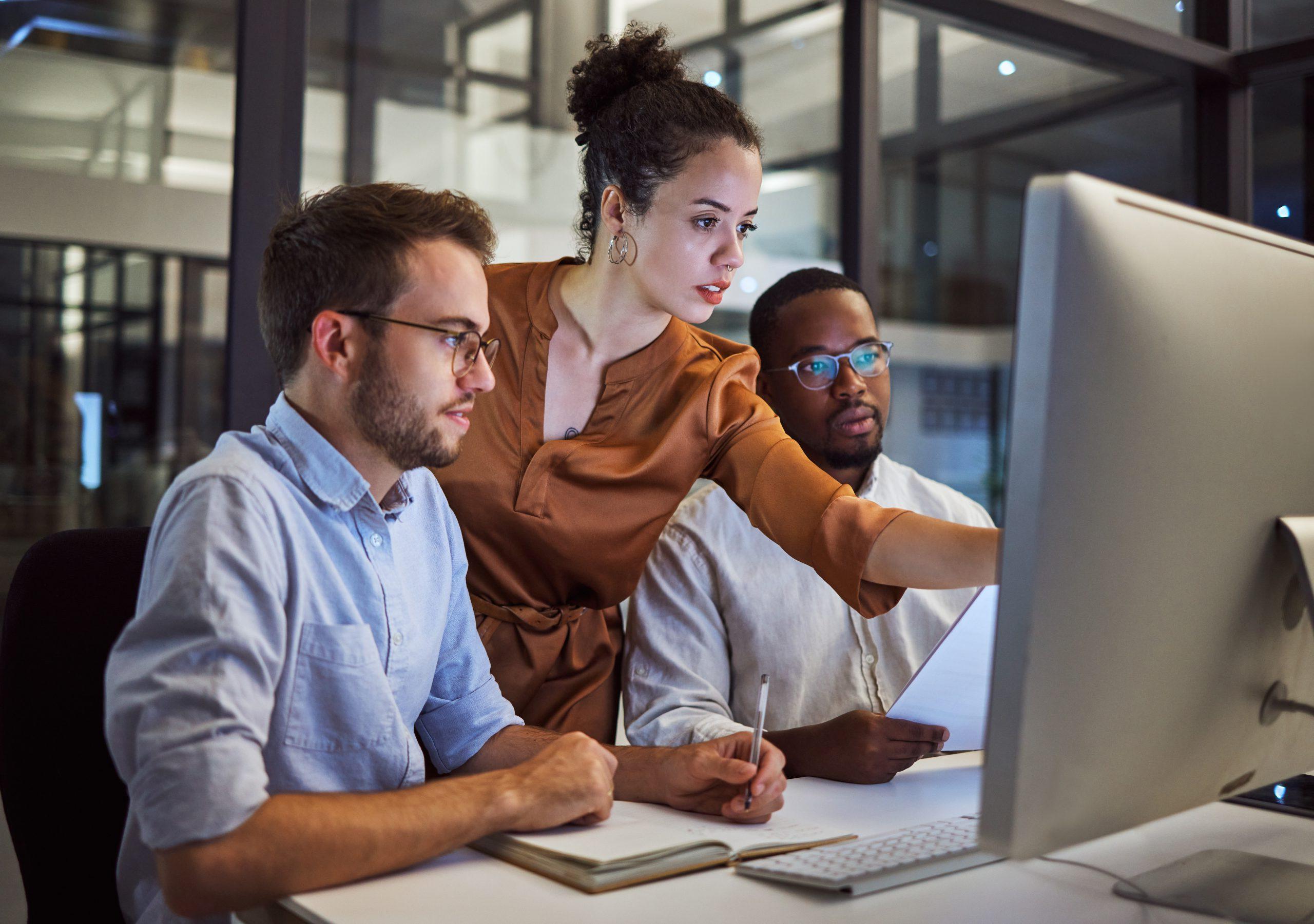 Three individual in front of a computer. One of them is pointing to the screen while the other two appear to be listening.