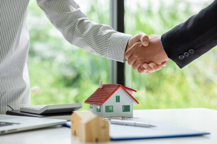 Two people shake hands, presumably over a real estate deal, 他们的手下面有一张桌子，上面有一个小的房屋模型.